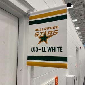 May include: A white banner with the text "MILLBROOK STARS" in orange and green, and a green star. Below, it reads "U13-LL WHITE." The banner is hanging on a wall near a door and a sign that says "Change Room 10."