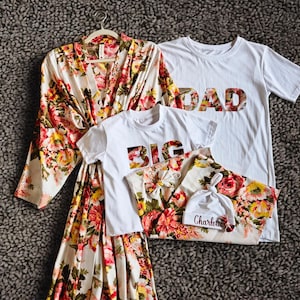 May include: A floral print robe, two white t-shirts with the words "DAD" and "BIG" in floral print, a matching floral print blanket, and a white hat with the name "Charlotte" on it. The items are arranged on a textured grey surface.