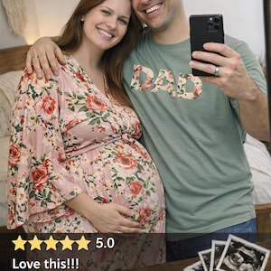 May include: A sage green short-sleeved t-shirt featuring the word DAD printed in a floral pattern with red roses and green leaves. The shirt is worn by a person, paired with a pink floral dress with a matching rose print and wide sleeves.