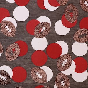 Könnte beinhalten: Draufsicht auf verstreute Konfetti in Rot, Weiß und glitzerndem Braun. Die Konfetti umfassen Kreise und American-Football-Formen, ideal für Sportpartys oder Dekorationen. Der Hintergrund ist eine dunkle Holzmaserung.