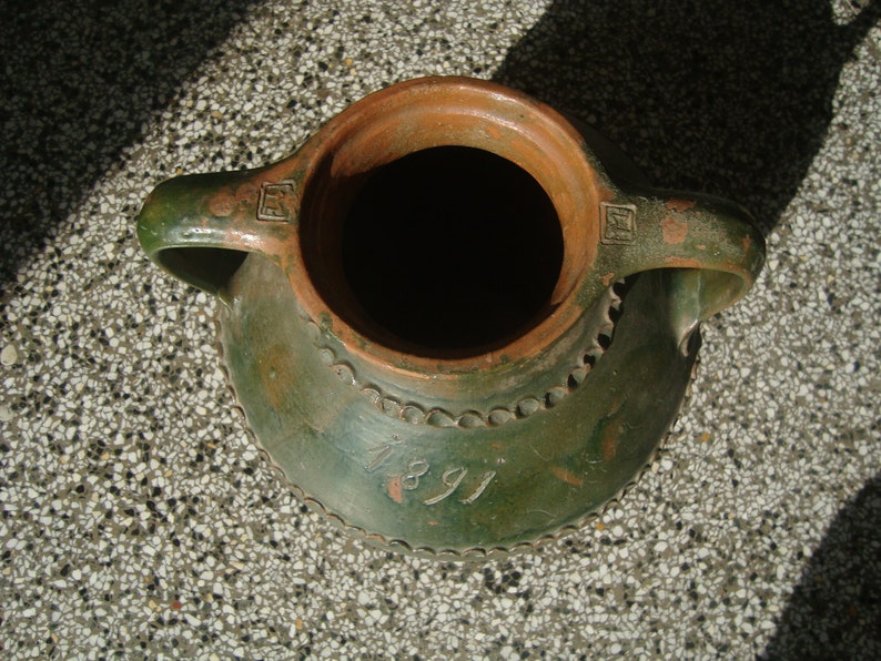 Peut inclure: Une cruche en c&eacute;ramique &eacute;maill&eacute;e verte avec deux anses et un int&eacute;rieur en terre cuite brune. La cruche pr&eacute;sente un motif d&eacute;coratif autour du bord et une date, "1891", inscrite sur le devant.