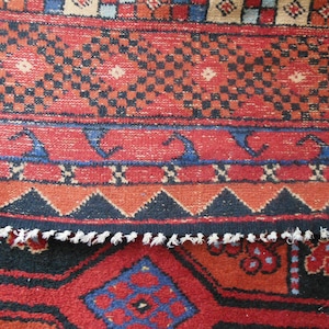 May include: Close-up of a red, blue, and black patterned rug with a fringed edge. The rug has a geometric design with repeating patterns.