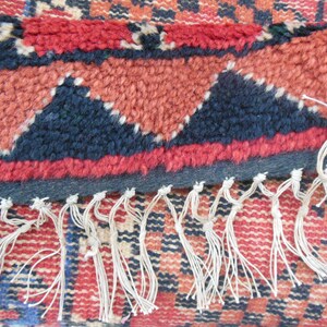 May include: Close-up of a red, black, and white woven rug with fringe. The rug has a geometric pattern with triangles and squares.