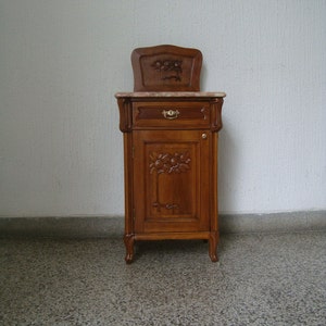 Può includere: Un piccolo comodino in legno con un cassetto e una porta dell'armadio. Il comodino ha un piano in marmo e dettagli floreali intagliati. Il comodino è di colore marrone chiaro.