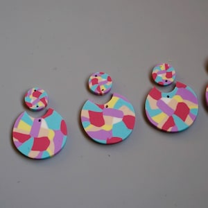 May include: A set of three pairs of colorful earrings. Each earring is made of two pieces of polymer clay, one round and one half-circle. The round pieces are solid, while the half-circle pieces have a hole in the center. The colors of the clay are pink, yellow, blue, and purple.