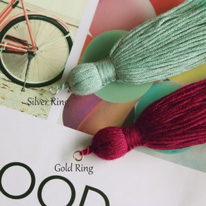 May include: Two decorative tassels, one in a soft seafoam green and the other in a deep burgundy. Each tassel has a rounded top and a long, flowing fringe. The seafoam green tassel has a silver ring, while the burgundy tassel has a gold ring.