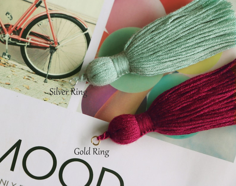May include: Two decorative tassels, one in a soft seafoam green and the other in a deep burgundy. Each tassel has a rounded top and a long, flowing fringe. The seafoam green tassel has a silver ring, while the burgundy tassel has a gold ring.