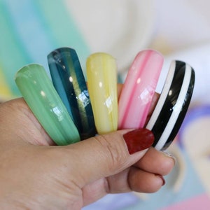 May include: A set of six colorful bangle bracelets. The bracelets are made of a translucent material and have a striped pattern. The colors of the bracelets are green, blue, yellow, pink, black, and white.