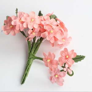 May include: A bundle of artificial pink flowers with yellow centers and green stems and leaves. The flowers are clustered together, with some individual blooms scattered around. The background is plain white.