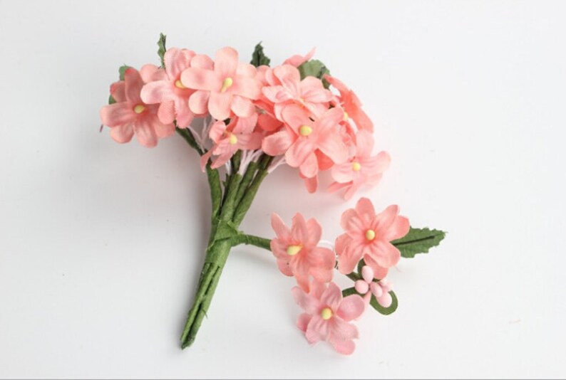 May include: A bundle of artificial pink flowers with yellow centers and green stems and leaves. The flowers are clustered together, with some individual blooms scattered around. The background is plain white.