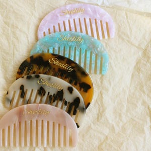 Può includere: Cinque pettini colorati per capelli nei toni del rosa, verde acqua, tartarugato e bianco con macchie nere. Ogni pettine ha una parte superiore curva e denti verticali. La parola "Shestily" è scritta in oro su ogni pettine.