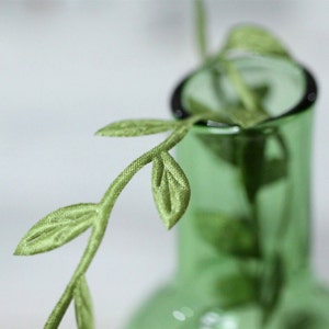 May include: A green glass vase with a single stem of green silk leaves.