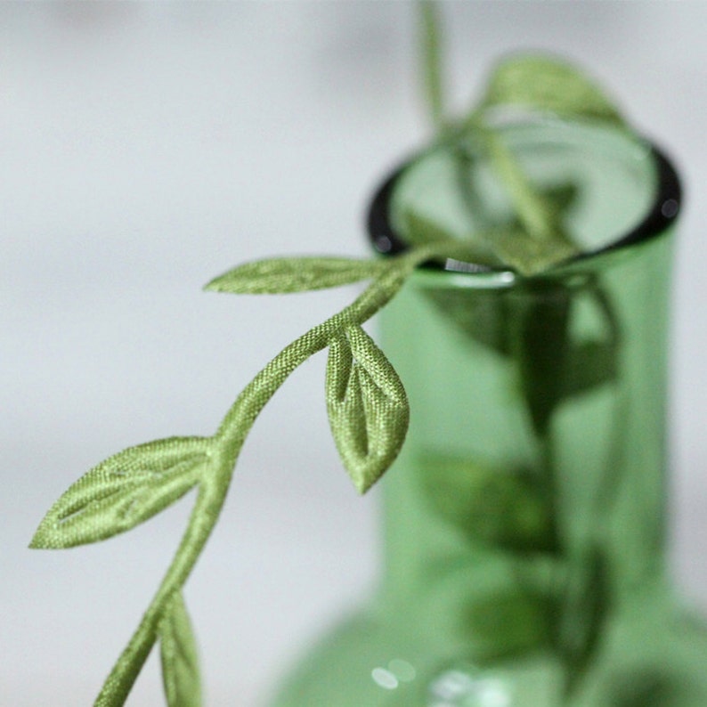 May include: A green glass vase with a single stem of green silk leaves.