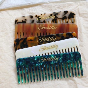 Puede incluir: Una pila de cinco peines en varios colores y patrones, incluyendo carey, marrón, blanco y verde. Cada peine tiene la palabra "Sheslily" en letras doradas. Los peines están colocados sobre una tela beige.