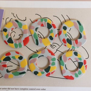 Könnte beinhalten: Sechs bunte, runde, keramische Ohrringe mit geometrischem Muster. Die Ohrringe sind in den Farben Rosa, Grün, Gelb, Rot und Blau.