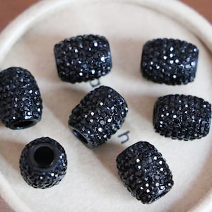 May include: Seven black rhinestone-covered barrel-shaped beads. The beads are all the same size and shape, and they are arranged in a circle on a white surface.