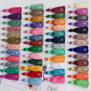 May include: A collection of 50 colorful tassels, each with a loop for hanging. The tassels are arranged in rows, showcasing a wide range of colors including pink, purple, green, orange, blue, and neutral tones. Each tassel is numbered for easy identification.