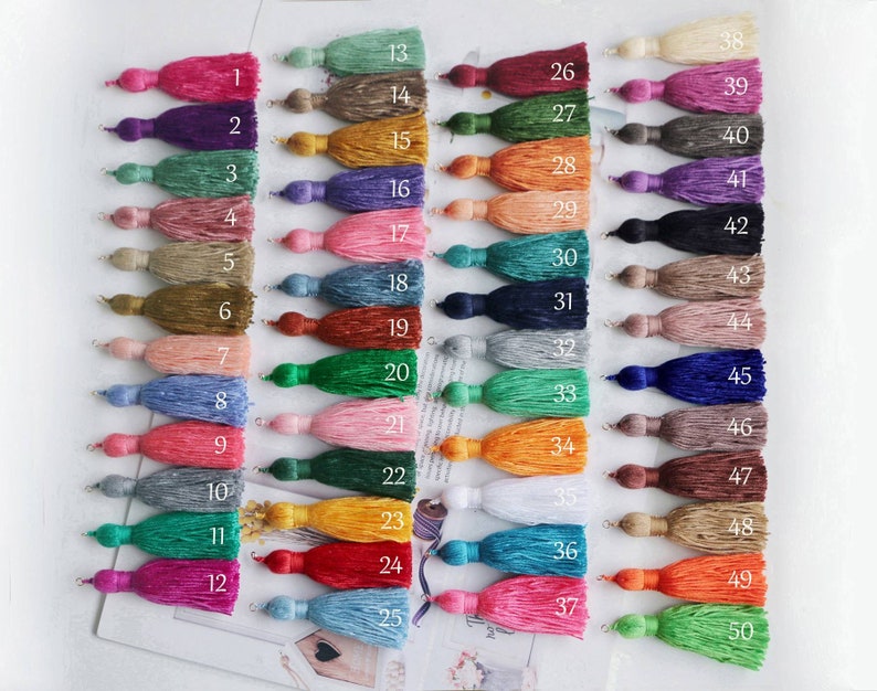 May include: A collection of 50 colorful tassels, each with a loop for hanging. The tassels are arranged in rows, showcasing a wide range of colors including pink, purple, green, orange, blue, and neutral tones. Each tassel is numbered for easy identification.