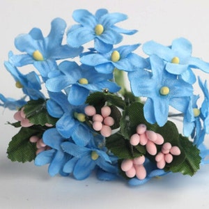 May include: A cluster of artificial flowers featuring light blue blossoms with yellow centers. The arrangement includes green leaves and small pink berry-like accents, creating a delicate and colorful floral display.