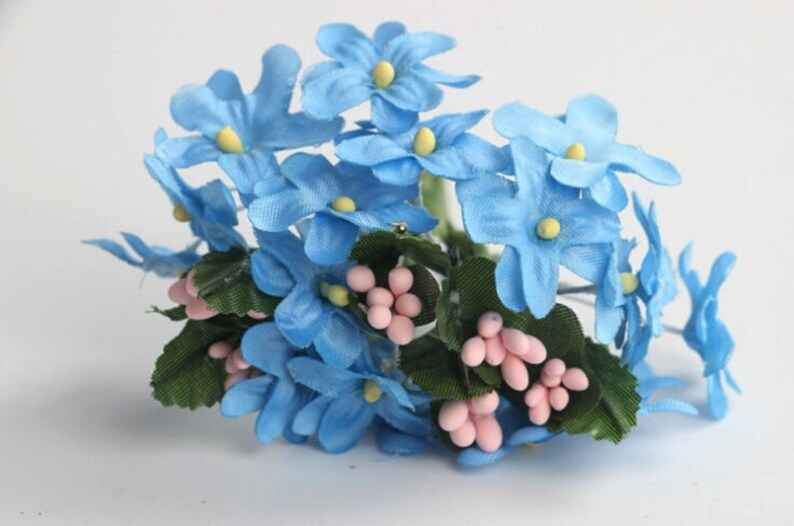 May include: A cluster of artificial flowers featuring light blue blossoms with yellow centers. The arrangement includes green leaves and small pink berry-like accents, creating a delicate and colorful floral display.