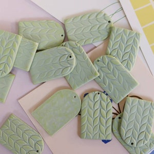 May include: A collection of green ceramic leaf-shaped tags with two holes each. The tags are arranged on a light pink surface.