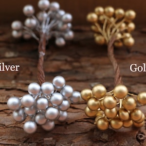 May include: Two sprigs of faux berries, one silver and one gold, on a brown wooden background. The sprigs are made of wire and have small, round berries.
