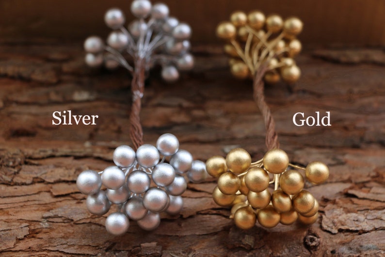 May include: Two sprigs of faux berries, one silver and one gold, on a brown wooden background. The sprigs are made of wire and have small, round berries.