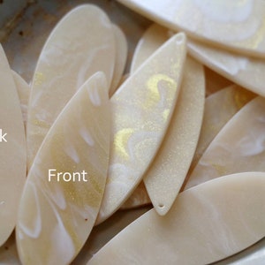 May include: A close-up of a pile of cream-colored resin leaf-shaped pendants with gold glitter and white swirls. The back of one pendant is plain cream-colored. The text "Back" and "Front" is written on the pendants.