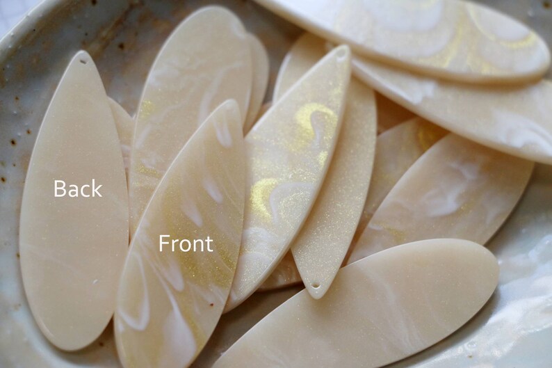 May include: A close-up of a pile of cream-colored resin leaf-shaped pendants with gold glitter and white swirls. The back of one pendant is plain cream-colored. The text "Back" and "Front" is written on the pendants.