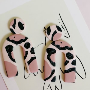 May include: A pair of pink and black statement earrings with a geometric arch shape and a smaller circle design. The earrings have a black and pink animal print pattern.