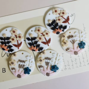 Puede incluir: Cinco adornos redondos de arcilla blanca con diferentes diseños florales. Los diseños presentan flores, hojas y tallos en tonos marrones, beige, rosa y azul verdoso.