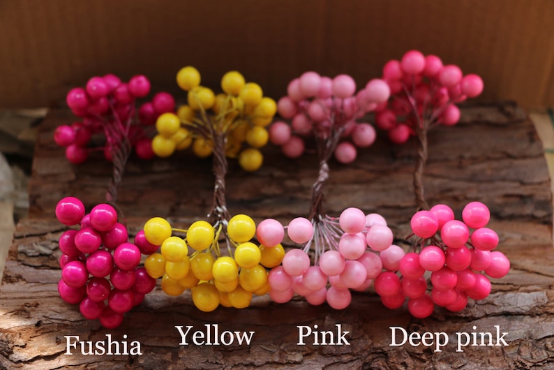May include: Five sprigs of faux berries in various shades of pink and yellow. The sprigs are arranged in a row on a wooden surface. The colors are labeled "Fuschia", "Yellow", "Pink", and "Deep pink".