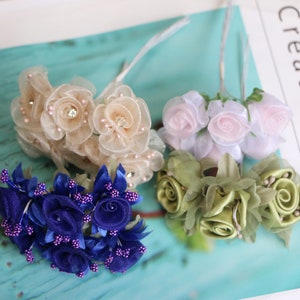 May include: Four bunches of artificial flowers, each with a different color and style. The first bunch is beige with a pearl-like center, the second is light pink, the third is dark blue with purple accents, and the fourth is green.