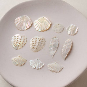 Op de afbeelding: Een verzameling witte, iriserende schelp- en blaadjesvormige bedels. De bedels hebben een gaatje bovenaan om op te hangen.