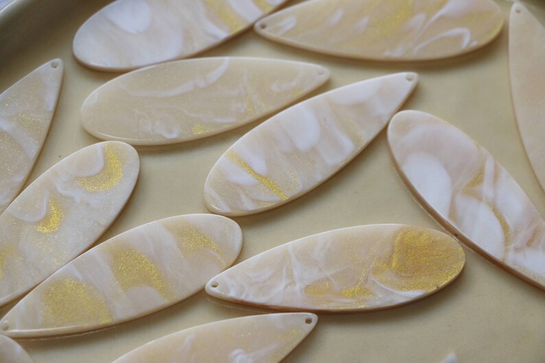 May include: A collection of oval-shaped resin blanks with a marbled pattern in white and gold. The blanks are perfect for crafting earrings, pendants, or other jewelry.