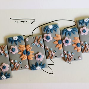 May include: Rectangular gray decorative tiles with floral designs. Each tile features a different arrangement of flowers in shades of orange, blue, and pink, with small blue and pink berry accents. The tiles are arranged in a slightly curved line.