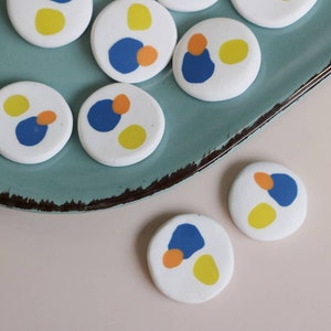 May include: A set of round, white ceramic coasters with abstract designs in blue, yellow, and orange. The coasters are arranged on a teal plate.