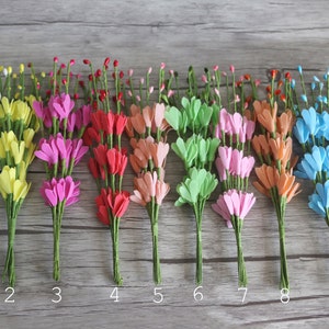 May include: Ten bunches of artificial flowers in various colours, including white, yellow, pink, red, orange, green, light pink, light blue, and purple. The flowers are arranged in a row on a wooden surface.