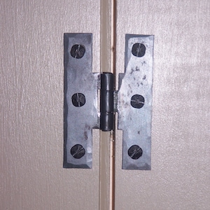 Puede incluir: Una bisagra de metal negro con cuatro tornillos visibles a cada lado. La bisagra está unida a una puerta de madera marrón claro.