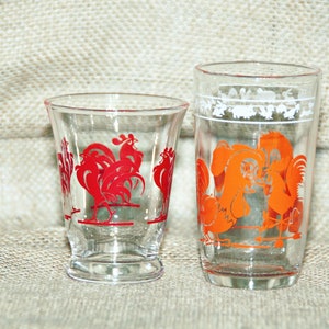 May include: Two clear glass drinking glasses with rooster designs. The smaller glass has red roosters and the taller glass has orange roosters.