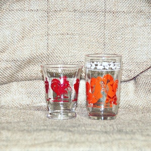 May include: Two clear glass drinking glasses with red and orange rooster designs. The glasses are on a burlap background.