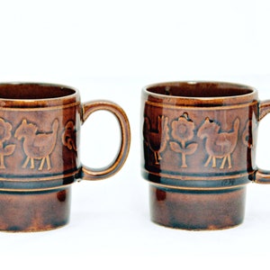 May include: Two brown ceramic mugs with a stacked design. Each mug features a repeating pattern of a rooster, flower, and horse.