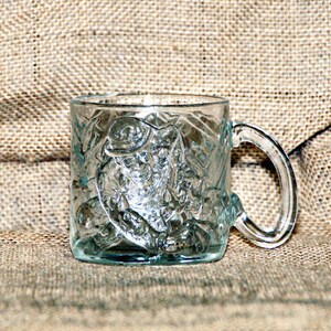 May include: A clear glass mug with a raised design of a bat and the words "Batman" on the side. The mug has a handle and is sitting on a burlap surface.