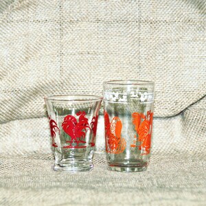 May include: Two clear glass drinking glasses with rooster designs. The smaller glass has a red rooster design and the taller glass has an orange rooster design.