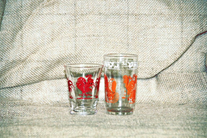 May include: Two clear glass drinking glasses with rooster designs. The smaller glass has a red rooster design and the taller glass has an orange rooster design.
