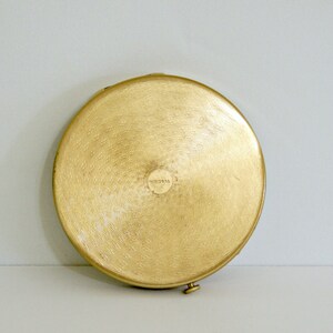 May include: A round, gold-colored compact with a slightly raised, circular design in the center. The compact has a small, round, gold-colored latch on the side.