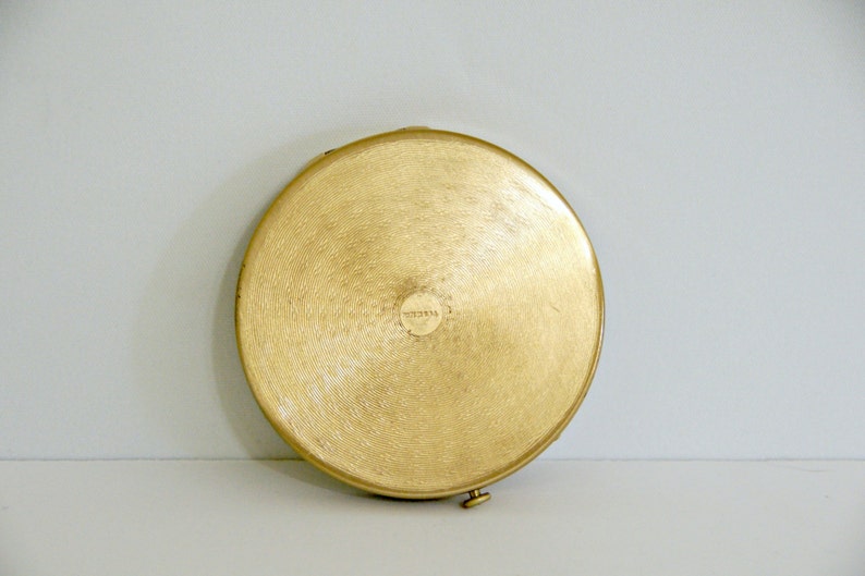 May include: A round, gold-colored compact with a slightly raised, circular design in the center. The compact has a small, round, gold-colored latch on the side.