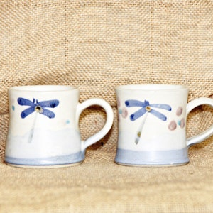 Könnte beinhalten: Zwei weiße Keramikbecher mit einem blauen Libellen-Design. Die Becher haben einen hellblauen Boden und Henkel. Die Libelle ist blau mit einem gelben Zentrum bemalt. Auf den Bechern sind auch rosa und blaue Punkte gemalt.