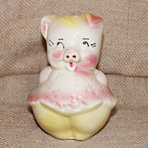 May include: A ceramic piggy bank with a pink and yellow dress. The pig has a white face with black eyes and a pink nose.