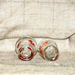 May include: Two clear glass shot glasses with red rooster designs. The glasses are upside down on a burlap surface.
